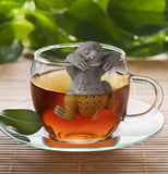 The gray smiling sloth is hanging from the rim of a clear glass filled with tea that's sitting the table. 