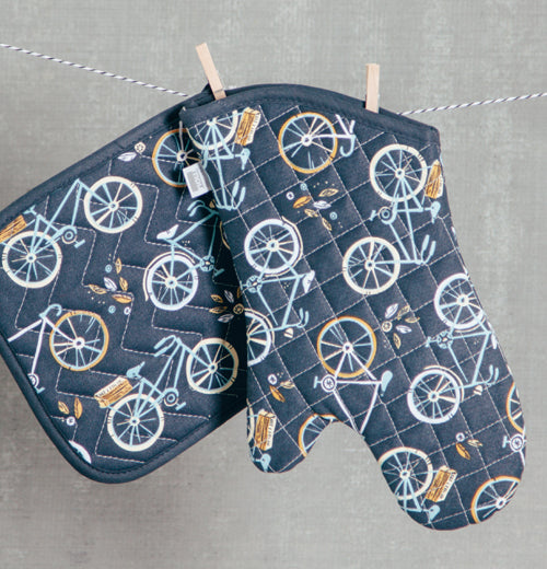 The navy blue oven mitt with white and golden bicycle artwork is hanging on a line next to a potholder with the same color and design.
