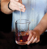 A man's hands are shown dipping a long stirring spoon into the liquid inside the crisscross patterned glass.