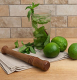 The wood muddler is shown lying on a wooden table next to some limes, and a drink full of leaves.