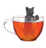 The black cat shaped infuser with its paws hanging over the edge of a clear glass cup it sits in, the cup is filled up with tea.