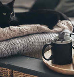 A picture of a black cat on some blankets with the cat shaped infuser on the side of a cup.