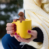 Woman holding a yellow mug in a yellow sweater and blue pants with the top of a red dog shaped tea infuser hanging on the rim of the mug.