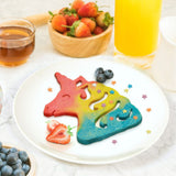 Red, yellow, and blue colored unicorn head shaped pancake covered with star sprinkles on round white plate with blueberries and sliced strawberries laying next to them. A bowl of blueberries is off to the lower left and on top is a clear cup of syrup, a bowl of strawberries, and a glass of orange juice.