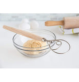 "Danish Dough" whisk with wooden handle on clear glass bowl with dough in it on white table with eggs and rolling pin on yellow striped white towel.