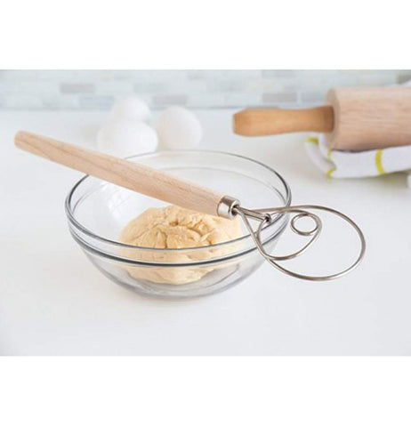 "Danish Dough" whisk with wooden handle on clear glass bowl with dough in it on white table with eggs and rolling pin on yellow striped white towel.