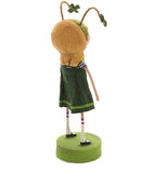 A polyresin figurine of a young, blonde-haired girl. She wears a green and gold painted four-leaf clover headband, a green dress, white socks with two green stripes at the top, and Mary Jane shoes. She is standing angled facing the right, with her back visible.