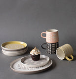 Side plates stacked up and white and gray dinner plate with chocolate cupcake on top of gray and white side plate. There are three mugs next to all the plates on a dark gray background.