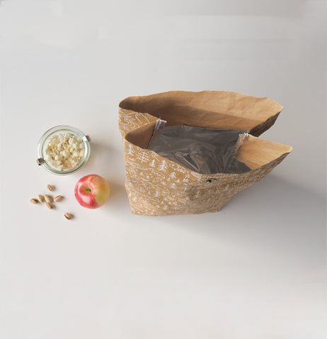 Open brown lunch bag with white outdoor design and silver insulation next to a red and yellow apple, sunflower seeds, and cup of popcorn next to it.