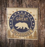 The box with a navy blue and cream colored circular bear symbol with the words, "The Great Outdoors" In navy blue and cream colored lettering and pine trees below the words sits on a wooden surface.