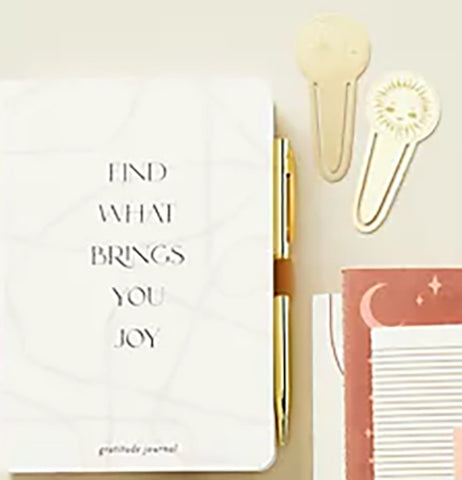 Two metal page tabs (one with a half moon, the other with a sun) next to a black and white journal and black, red, and white notepads. The journal cover reads "Find what brings you joy - gratitude journal."