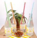 Four different colored reusable straws, one green, another orange, another red, and the last blue, being used in four sparkling water bottles on a table with nuts and a plant in the background