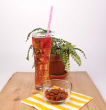 Its a pink reusable straw in a iced tea glass sitting on a table along with a bowl of nuts sitting on a white and yellow napkin with a plant in the background.