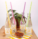 Four different colored reusable straws, one yellow, another pink, another purple, and the last lime green, being used in four sparkling water bottles on a table with nuts and a plant in the background