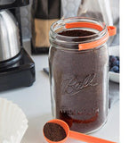 Orange coffe spoon filled with coffee on table next to coffee filter with mason jar filled with coffee with orange clip on it.
