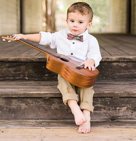 A brown haired little boy wearing the green bow tie with pink roses around his neck sits while he plays with small guitar.