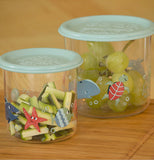 Two clear snack containers with Ocean themed sea life in red and different shades of blue sitting on a wood counter. One has green grapes in it, the other has cut up zucchini sticks. Each snack containers has a light blue lid attached.