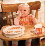 A baby is shown wearing the princess bib with a plate and cup of a similar design on the high chair.