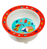 A bowl is seen from a downward angle. The rim is red, and has small designs on it like trees, compasses, and lanterns. The sides of the bowl are white. The bottom of it has an anthropomorphic red dog with black ears wearing a light brown top and roasting a hot dog over a campfire. To the right of the dog, there is a tent. There are five trees in the background.