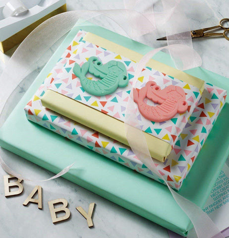 Both colored, teal and salmon pink colored teether, sit on a polka dot patterned paper wrapped around a teal green box.