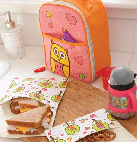 The pink and orange lunch box with the owl and hearts on it is shown sitting on a table in front of some bags with a sandwich and some pretzels. A matching cup on the right of the lunch box is shown.