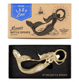 The brown box lid with the blue ribbon and the black picture of a mermaid and sailboat is shown at the top of the image. At the bottom is the black casing in which the brass mermaid bottle opener comes.