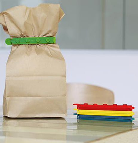 Three of the "Stackable Bag Clips" are on the table with the green one clipping the paper lunch bag closed . 