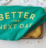 Green cotton, polyurethane, and polyester baking dish cover saying "Better the Next Day" in yellow with corner pulled up on lower right of baking pan.