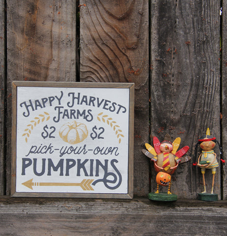 a box sign with happy harvest farms, pick-your-own, and Pumpkins sitting against a wood background with two figurines, on of a kid in a turkey costume and the other of a girl in a native american costume.