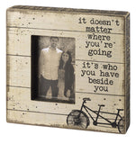 An off-white wooden box sign with the phrase "It doesn't matter where you're going it's who you have beside you" above a two person bicycle