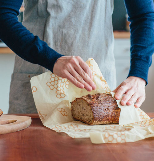 A person rewrapping yellow Food Wrap around the cut loaf of bread on a brown table.