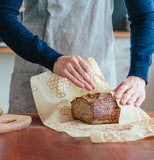 A person rewrapping yellow Food Wrap around the cut loaf of bread on a brown table.