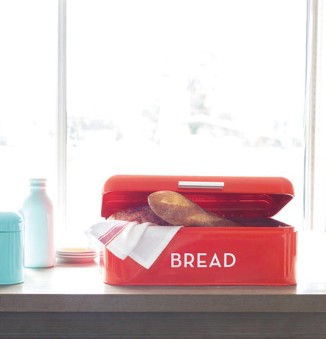 A red bread box with the white words Bread on it is sitting on a wooden table with a dish towel and beget poking out of its open lid