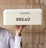 A Cream Colored Bread Box with the black lettering Bread is being held in the air by someone in a white cross stitch shirt