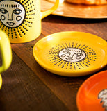 The yellow coaster with a white faced sun in the center is shown sitting on a wooden table next to a mug with the same design.