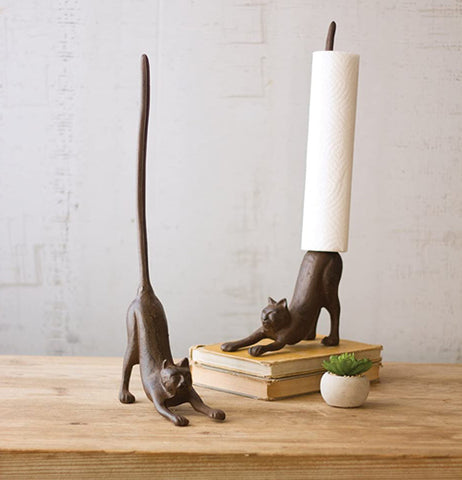 A pair of cast-iron cat paper towel holders one placed on stacked books with white paper towel roll facing left with one facing right without a towel roll both on a natural wooden table.