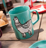 The turquoise ceramic mug with a llama is shown next to a coaster with the same design sitting on a wooden table.