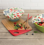 This red rectangular cutting board with a red dipped edge sitting on a table with a blue bowl with the berry patch bowl cover sitting on top of it along with red dipped wooden salad servers, a green bowl with the berry patch bowl cover half off is sitting to the right