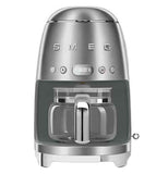 A stainless steel coffee machine with a coffee carafe set up inside it. The base has small "feet" that elevate it slightly. The brand name "Smeg" and buttons are across the body.
