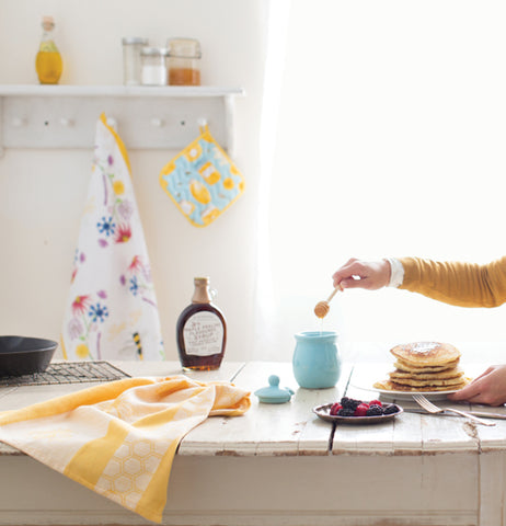 The yellow honeybee dishtowel is shown sitting on a table with syrup, honey, and pancakes.