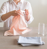 Someone using an orange and white striped dishcloth to clean a glass with two blue and white striped dish towels sitting folded on the table in front along with another glass.