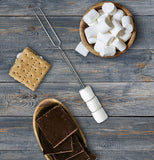 skewer laying on a wood patio alongside a bowl of marshmallows and graham crackers and someone's shoe poking in at the bottom.