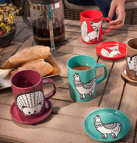 The teal llama mug and coaster are shown next to two red cups and coasters, one with a hedgehog design, the other with a fox design.