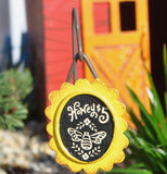 A close up fo the honey sign garden pick that says "honey $5" above a bee in black and white. There is a small glimpse of a red barn with an orange door in the background.