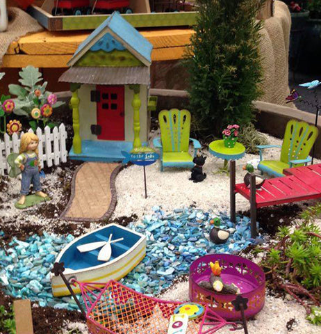 A lake themed mini garden features a little hut next to a green and blue table set, with a little pebble river running down the middle with a boat sitting on it and the a red dock to the right, a red hamuk and pink campfire are at the bottom of the garden
