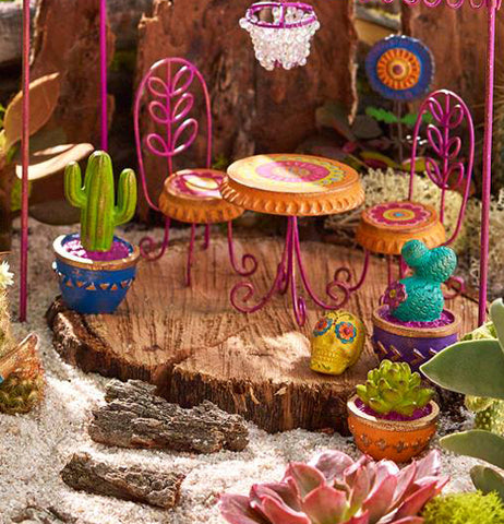 A desert themed mini garden that features The (Mini) Cactuses in orange, purple, and blue pots, a pink floral patio set and other mini garden ornaments sitting on top of a piece of wood. 