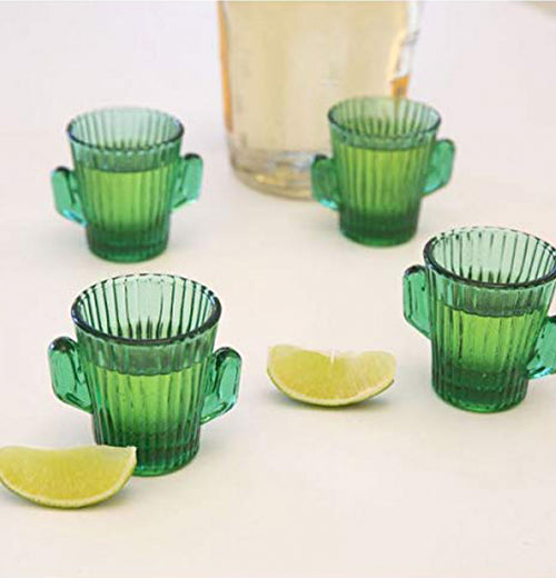 Four cactus-shaped shot glasses are shown sitting on a table next to a large glass pitcher, and two lime wedges.