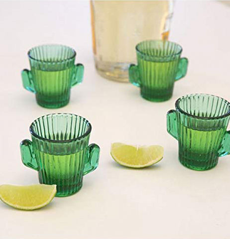 Four cactus-shaped shot glasses are shown sitting on a table next to a large glass pitcher, and two lime wedges.