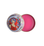 A circular red, blue, and gold art neaveau tin has pink lip balm in it.