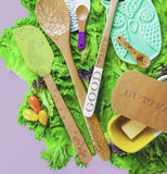 The green spatula with the hedgehog picture on its head is shown mixed with other spoons and spoon holders all sitting on grass.